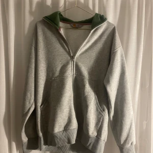 Grå hoodie från Carhartt - Säljer en grå hoodie från Carhartt med dragkedja och en mysig grön insida i luvan. Den har en klassisk passform med ribbade muddar och fickor framtill. Superbekväm och stilren!