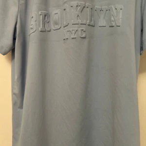 Ljusblå t-shirt med Brooklyn-tryck - Säljer en ljusblå t-shirt med texten 'BROOKLYN NYC' i relief på framsidan. T-shirten har en normal passform och är kortärmad, perfekt för en avslappnad stil. Materialet känns mjukt och skönt mot huden.