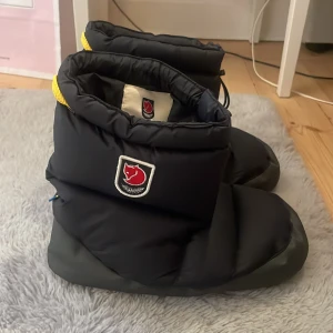 Svarta Fjällräven Expedition tofflor - Säljer ett par svarta Fjällräven Expedition tofflor i storlek S. 👌💕De är supervarma och perfekta för vintern. Tofflorna har en praktisk dragsko för att justera passformen och en snygg  fjällräven detalj!💕 Perfekta för kalla dagar hemma eller i stugan! Det är väldigt bra skick har bara används 1-3 ggr, därför är det högt pris. Nypris runt 1300-1600kr. 💕👌Tveka inte om frågor!💕 Priset kan justeras!❣️