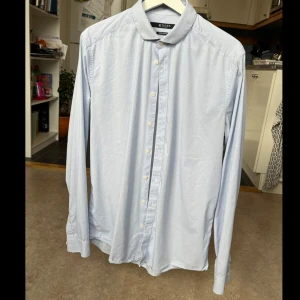 Tiger of Sweden skjorta  - Ljusblårandig skjorta, syns bäst på bild 3-5. Litet märke längst ner på ryggen, syns inte om skjortan är instoppad, bild 5. Sparsamt använd, säljes då den inte passar. Storlek L/40-41 i slim fit stretch, material 63% bomull, 32% polyamid, 5% elastan.