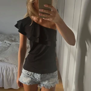Svart oneshoulder topp - Snygg svart oneshoulder topp med volangdetaljer. Toppen har en tight passform och är tillverkad i ett mjukt och bekvämt material.
