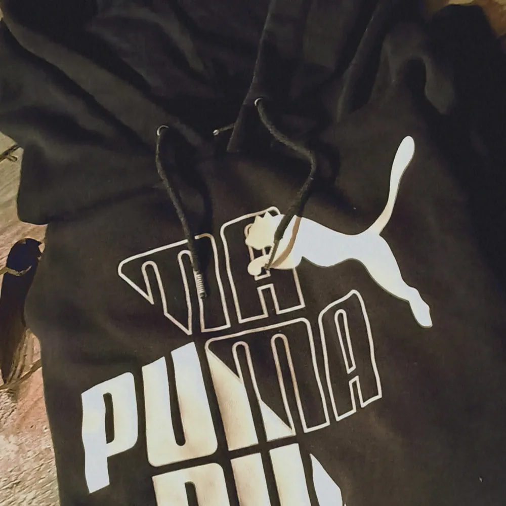 Säljer en snygg svart hoodie från Puma. Den har en stor vit logga på framsidan och justerbara snören i huvan. Perfekt för en avslappnad stil eller träning. Passar bra till både jeans och träningsbyxor!. Tröjor & Koftor.