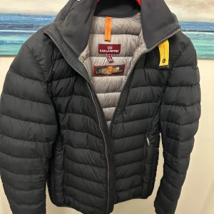 Svart dunjacka från Parajumpers - Säljer en snygg och lätt svart dunjacka från Parajumpers. Jackan har en cool gul detalj vid dragkedjan och en broderad patch på ärmen. Perfekt för höst och vinter med sin varma och quiltade design. Den har en normal passform och är superbekväm att ha på sig. Passar både till vardags och lite mer uppklädda tillfällen.