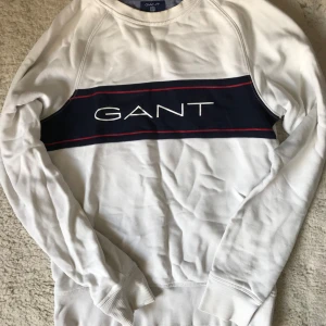 Vit tröja från GANT - Säljer en stilren vit tröja från GANT med ett blått band och röd detalj över bröstet där märkesnamnet är tryckt. Tröjan har långa ärmar och ribbade muddar vid ärmslut och nederkant. Perfekt för en avslappnad stil.