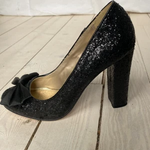 Svarta glittriga pumps med rosett - Svarta glitterpumps från Ellos med en sidenrosett framtill. Skorna har 10 cm klack och piffar till vilken outfit som helst. Storlek 38 men är lite smala framtill. Kan nog också passa en 37a. Använda vid ett tillfälle.  