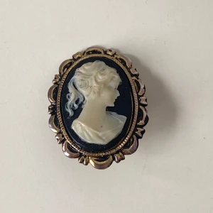  brosch med cameo-motiv - Elegant  brosch med en klassisk cameo-design. Broschen har en vacker relief av en kvinnas profil i vitt mot en svart bakgrund, inramad av en dekorativ guldfärgad kant. Perfekt för att ge en tidlös touch till din outfit.