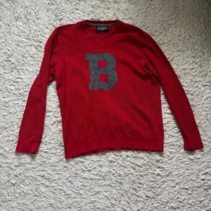Röd tröja med bokstav från Bondelid - Säljer en snygg röd tröja från Bondelid med en stor grå bokstav 'B' på framsidan. Tröjan har långa ärmar och en klassisk rund halsringning. Perfekt för en avslappnad stil.