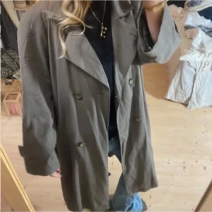 Olivfärgad trenchcoat - Snygg olivfärgad trenchcoat med knappar framtill. Perfekt för lager-på-lager-stil och ger en klassisk look. Passar bra till jeans och en enkel topp.