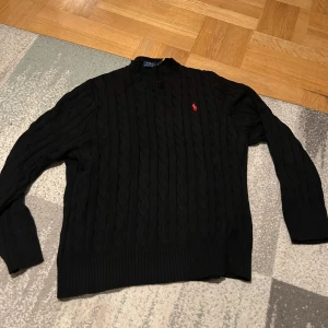 Polo Ralph Lauren stickad tröja - Säljer en stilren svart kabelstickad tröja från Polo Ralph Lauren. Tröjan har en klassisk design med ett rött broderat märke på bröstet. Perfekt för kyligare dagar med sin mjuka och bekväma passform.