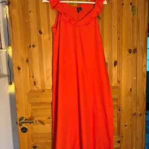 Röd klänning med volangdetalj - Säljer en härlig orange klänning med volangdetalj vid halsringningen. Klänningen har en avslappnad passform och är perfekt för varma dagar. Den är ärmlös och har en enkel design som gör den lätt att styla.