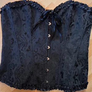 Fin korsett i gotisk stil! - Jättefin korsett i gotisk stil med hakknäppning och mjuka skenor fram i svart broderad spets och satin med rygglapp och snörning bak som kan justeras flera dm. Mått: H: 40 cm, 85 cm runtom på innersta snörning med utrymme för extra 20 cm. Perfekt skick.