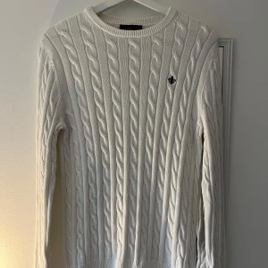 Vit stickad tröja från Morris - Passar 175-185 Säljer en stilren vit stickad tröja från Morris med kabelstickat mönster. Tröjan har en rund halsringning och ett diskret emblem på bröstet. Perfekt för kyligare dagar! 