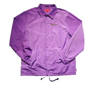 Lila jacka från Supreme - Säljer en snygg lila jacka från Supreme med knappar och broderad logga på bröstet. Jackan har långa ärmar och dragsko i nederkant för justerbar passform. Perfekt för en stilren look!