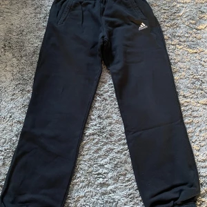 Svarta mjukisbyxor från Adidas - Säljer ett par svarta mjukisbyxor från Adidas med elastisk midja och sidofickor. Byxorna har en liten Adidas-logga på ena benet och är perfekta för en avslappnad stil. De är tillverkade i mjukt material för extra komfort.