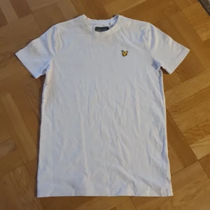 Vit t-shirt från Lyle & Scott - Säljer en klassisk vit t-shirt från Lyle & Scott med korta ärmar och en gul broderad logotyp på bröstet. Perfekt för en stilren och avslappnad look. Passar ungdomar i åldern 12-13 år. 