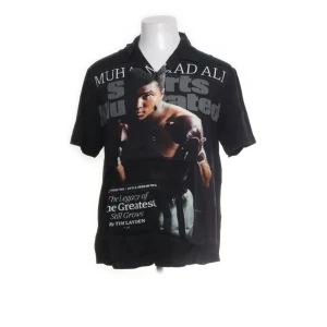 Svart kortärmad skjorta med Muhammad Ali-tryck - Cool svart skjorta från Sports Illustrated med stort tryck av Muhammad Ali på framsidan. Skjortan är kortärmad och har en avslappnad passform. Perfekt för dig som vill ha en unik och sportig stil.