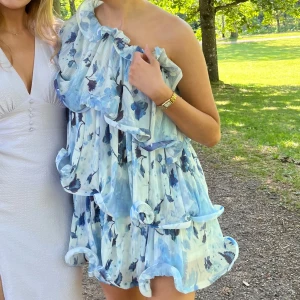 Blå blommig oneshoulder klänning från NAKD - Säljer en superfin blå blommig oneshoulder klänning med volanger. Perfekt för sommarens alla tillfällen. Klänningen har en luftig passform och är kort i modellen. Den har ett härligt mönster med olika blå nyanser och vita detaljer.