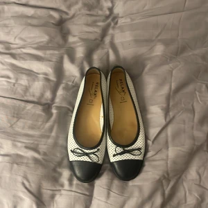 Svartvita ballerinaskor från Relax - Snygga svartvita ballerinaskor från Relax med prickigt mönster och dekorativ rosett. Skorna har en svart spetsig tå och är tillverkade i syntetmaterial. Perfekta för en stilren look.