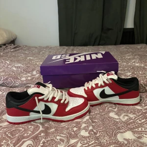 NIKE SB DUNK LOW PRO - VARSITY RED/BLACK-WHITE - INGÅR SVARTA SNÖREN! Snygga Nike Dunk Low sneakers i en klassisk färgkombination av rött, vitt och svart. Skorna har en ikonisk Nike-logga på sidan och kommer med vit snörning. Perfekta för en stilren look. 