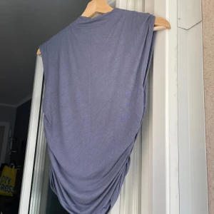 Grå och blå/grå ärmlösa toppar från Gina Tricot - Säljer två stilrena grå och grå/blå ärmlösa toppar från Gina Tricot. Topparna har en draperad design på sidorna som ger en snygg detalj. Endast använt vardera en gång och kommer tyvärr inte till användning längre.