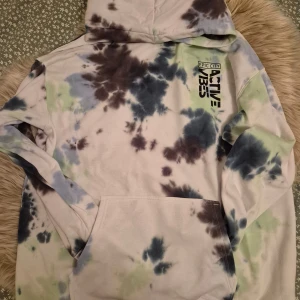 Tie-dye hoodie från Epic City - Säljer en cool tie-dye hoodie från Epic City med texten 'ACTIVE 135' på bröstet. Tröjan har en känguruficka och en bekväm huva. Färgerna är en mix av blått, grönt och brunt på en ljus bas.