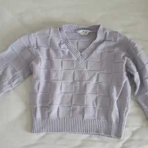 Ljuslila stickad tröja från H&M - Säljer en ljuslila stickad tröja från H&M med ett rutigt mönster. Tröjan har en v-ringad hals och långa ärmar. Perfekt för en stilren och bekväm look.