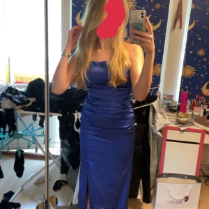 Blå långklänning med öppen rygg - Snygg blå långklänning i satin med en slits framtill. Klänningen har en öppen rygg med snörning och smala axelband som korsas i ryggen. (Använd en gång)