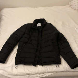 Svart pufferjacka från Zara - Snygg svart pufferjacka från Zara med dragkedja och justerbara snören i nederkant. Perfekt för kyligare dagar med sin vadderade design och långa ärmar. En stilren jacka som håller dig varm och trendig.
