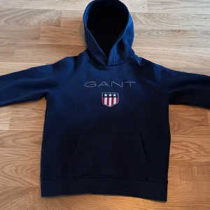 Mörkblå hoodie från GANT - Säljer en snygg mörkblå hoodie från GANT med ett broderat märke och logga på framsidan. Tröjan har en klassisk känguruficka och en bekväm huva. Tröjan är bara änvänd några gånger och är i ett väldigt bra skick. Perfekt för en avslappnad stil. Fråga gärna om fler bilder. Priset kan diskuteras. Nypris 600