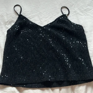 Svart glittrig linnetopp - Snygg svart linnetopp med tunna axelband och glittriga paljetter. Perfekt för en kväll ute eller festliga tillfällen. Toppen har en elegant och stilren design.