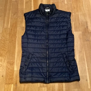 Mörkblå dunväst från McKinley - Säljer en mörkblå dunväst från McKinley. Västen har en dragkedja framtill och två fickor med knappar. Perfekt för kyligare dagar med sin quiltade design.