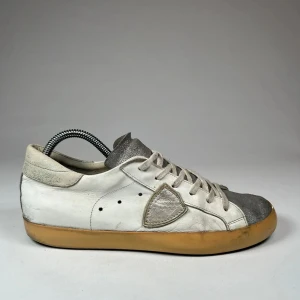 Vita och grå sneakers med snörning från fhilippe model - Snygga sneakers i vitt och grått med en unik design från fhilippe model. Skorna har en vit läderbas med grå detaljer på tån och plösen. De har en klassisk snörning och en gummisula i beige som ger en retro känsla.