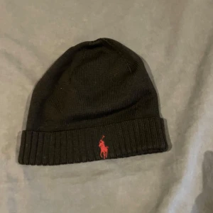 Svart mössa från Polo Ralph Lauren - Snygg svart mössa från Polo Ralph Lauren med röd broderad logga framtill. Mössan är ribbad och perfekt för kyliga dagar. En stilren accessoar som passar till det mesta.