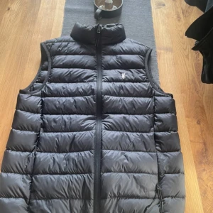 Svart dunväst från Polo Sport - Passar S/M                                                                          Från det populära märket polo Ralph Lauren.               Säljer en svart dunväst från Polo Sport med dragkedja och broderad logga på bröstet. Västen är perfekt nu till våren  och har en stilren design. Hör av er vid frågor//Albin