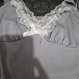 Grå pyjamas med spetsdetaljer - Säljer en söt grå pyjamas med vita spetsdetaljer och en liten rosett framtill. Toppen har smala axelband och byxorna har spetskant vid bensluten. Perfekt för mysiga kvällar hemma. Använd bara en gång. I storlek M