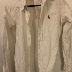 Vit skjorta från Ralph Lauren - Säljer en klassisk vit skjorta från Ralph Lauren med broderad logga på bröstet. Skjortan har långa ärmar och knappar framtill. Perfekt för en stilren look. Skjortan är i nyskick bara lite skrynklig! Storlek 6 som motsvarar S