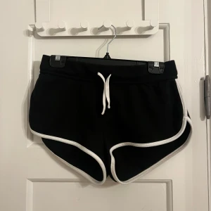 Svarta shorts med vita detaljer - Snygga svarta mjukisshorts med vita kanter och snörning i midjan. Perfekta för en avslappnad stil. De har en bakficka och är gjorda i ett mjukt material för extra komfort.