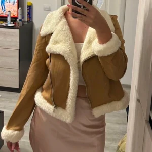 Beige mockajacka med fuskpäls - Snygg beige mockajacka med vit fuskpäls på krage, ärmslut och nederkant. Jackan har en dragkedja framtill och ger en trendig look. Perfekt för kyligare dagar när du vill vara både varm och stilren.