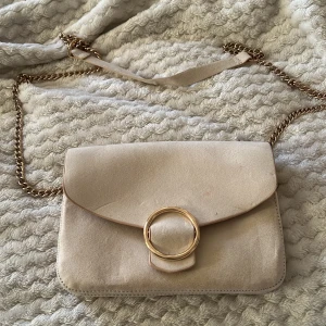 Beige axelväska med guldkedja - Snygg beige axelväska från H&M med en elegant guldkedja och en stor guldring på framsidan. Väskan har en praktisk knappstängning och är perfekt för att bära dina nödvändigheter med stil. Liten rosa prick mitt på väskan men ska försöka få bort den också, väskan är annars bara använd någon enstaka gång 