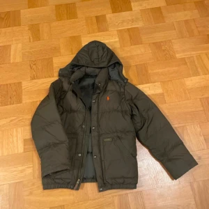 Ralph Lauren - Säljer en olivgrön dunjacka från Ralph Lauren med huva och dragkedja. Jackan har en broderad logotyp på bröstet och knappar framtill. Perfekt för kyliga dagar.