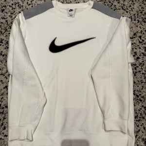 Vit sweatshirt från Nike - Säljer en stilren vit sweatshirt från Nike med grå axelpartier och det klassiska svarta Nike-logotypen på bröstet. Tröjan har långa ärmar och är perfekt för en sportig look.