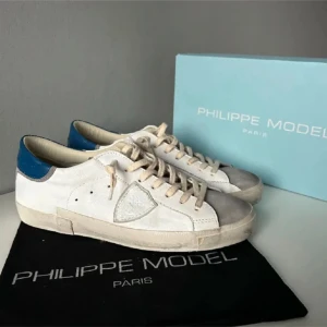 Philippe Model skor  - Stilrena vita sneakers från Philippe Model med blå detalj på hälen. Skorna har snörning och en diskret logga på sidan. Perfekta för en avslappnad look.