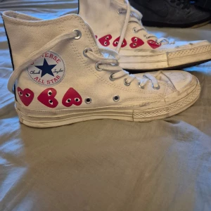 Vita Converse All Star med hjärtdetaljer - Säljer ett par vita Converse All Star sneakers med unika röda hjärtan med ögon på sidan. Skorna har klassisk snörning och den ikoniska Converse-loggan. Perfekta för en cool och avslappnad stil.Det är ett samarbete med comme des garcons. 