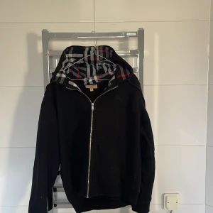 Svart hoodie från Burberry - Säljer en snygg svart hoodie från Burberry med dragkedja och en klassisk rutig insida i huvan. Den har ett broderat emblem på bröstet och långa ärmar. Perfekt för en stilren look.