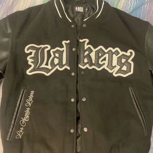 Svart Lakers bomberjacka - Säljer en svart bomberjacka med Lakers-logga. Jackan har långa ärmar i skinn och broderade detaljer på både fram- och baksida. Den har en klassisk bomberstil med ribbade muddar och krage.
