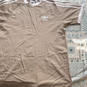  Adidas t shirt  - Säljer en snygg beige t-shirt från Adidas med klassiska vita ränder på axlarna och det ikoniska logotypen på bröstet. T-shirten har korta ärmar och en bekväm passform. Perfekt för en avslappnad stil.