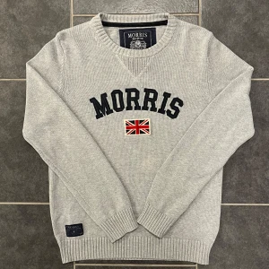 Grå tröja från Morris - Säljer en stilren grå tröja från Morris i 100% bomull. Tröjan har ett broderat Morris-märke och en brittisk flagga på framsidan. Den har en v-ringad hals och ribbade muddar vid ärmslut och nederkant.