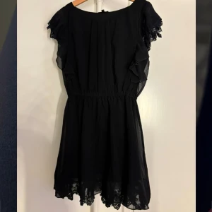 Svart klänning från KappAhl - Snygg svart klänning från KappAhl med volangdetaljer och spets vid ärmar och fåll. Perfekt för en stilren look. Klänningen har en elastisk midja för en bekväm passform.