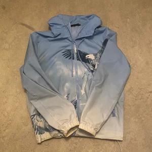 Blå vindjacka från Trapstar - Säljer en blå vindjacka från Trapstar med ett coolt örntryck på både fram- och baksidan. Jackan har dragkedja och långa ärmar, perfekt för blåsiga dagar. Den har en luva och elastiska muddar vid ärmsluten för extra komfort.