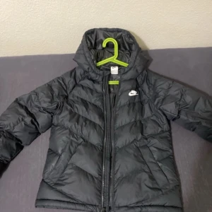 Svart dunjacka från Nike - Säljer en svart dunjacka från Nike med huva och dragkedja. Jackan har en stilren design med Nike-logga på bröstet. Perfekt för kyligare dagar. Köpt för 1500 säljer för 500. Pris kan diskuteras. Storlek L men funkar jätte bra för S och M. 